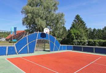 Buitenbasketbalveld met blauwe omheining en bomen op de achtergrond bij een glampingaccommodatie.
