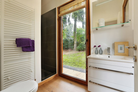 Modern tiny bathroom with large window overlooking greenery, sleek white vanity, shower, and towel radiator.
