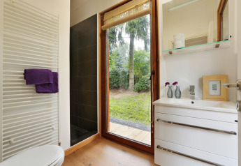 Modern kleine badkamer met grote glazen deur naar tuin, witte wastafel, inloopdouche en radiator.