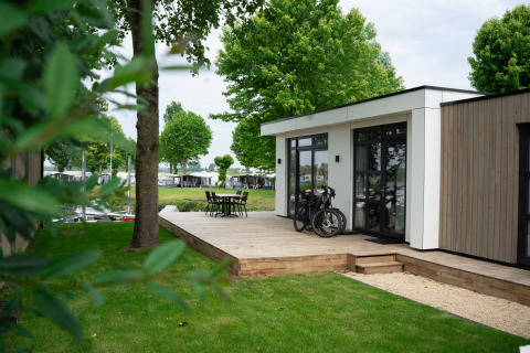 Lodge moderne nommé Villa Maritiem avec terrasse en bois, vélos et chaises de jardin, entouré d’arbres verts.