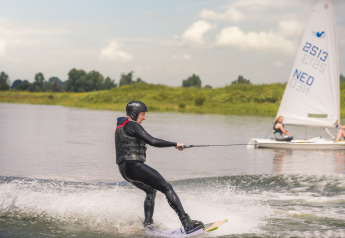 Una persona hace wakeboard en el lago junto a Villa Maritiem, con un velero visible al fondo.
