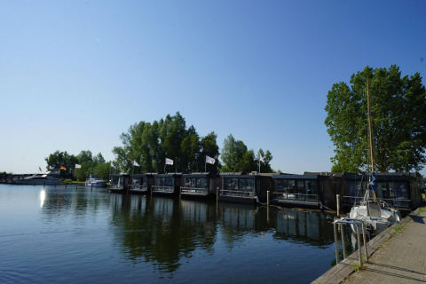 Zicht op woonboten aan een lodge aan het meer, omringd door bomen en boten, onder een blauwe hemel.