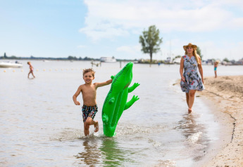 Niño corre con cocodrilo inflable verde en la playa, su madre lo sigue en Waterlodge 4, Zuiderzee, Países Bajos.