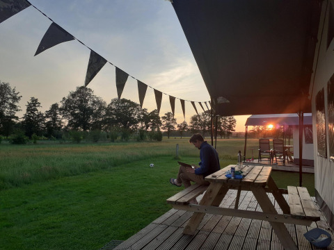 Man op picknicktafel bij glamping safaritent op De Regge-Vallei, Nederland, met zonsondergang op de achtergrond.