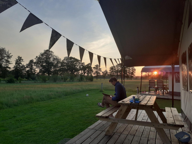 Man sits at a picnic table beside a glamping safari tent at De Regge-Vallei, Netherlands during sunset.