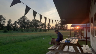 Mand sidder ved et picnicbord ved en glamping safari-teltplads i solnedgang på De Regge-Vallei, Holland.