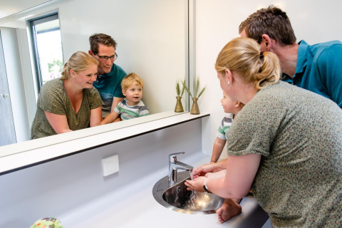 En familie vasker hænder sammen på et glamping sted, smilende foran spejlet ved en moderne vask.