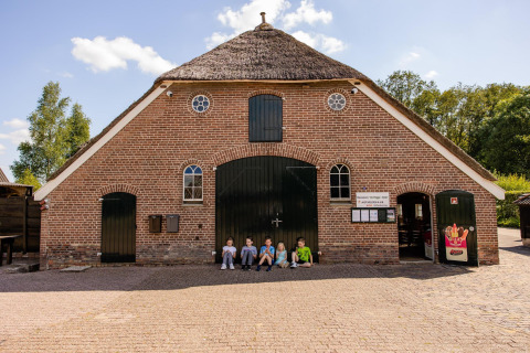Kinderen zitten voor een rustiek gebouw met rieten dak op een glamping onder een heldere hemel.