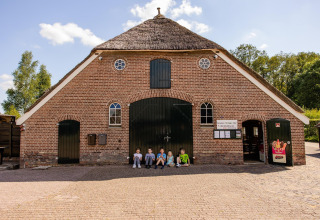 Kinderen zitten voor een rustiek gebouw met rieten dak op een glamping onder een heldere hemel.