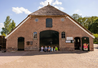 Kinderen zitten voor een rustiek gebouw met rieten dak op een glamping onder een heldere hemel.