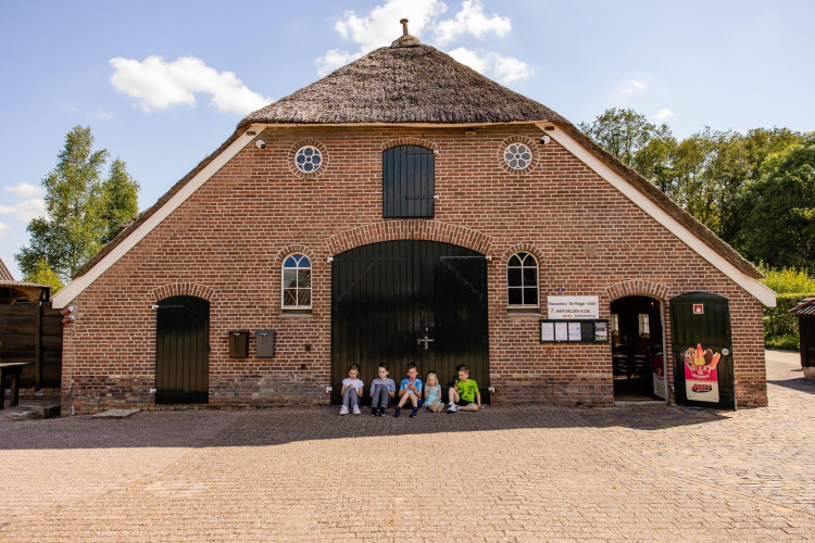 Children sit in front of a rustic thatched-roof building at a glamping site under a sunny sky.