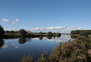 Zicht op een stille vijver met groen, helder water en blauwe lucht bij een glampingaccommodatie.