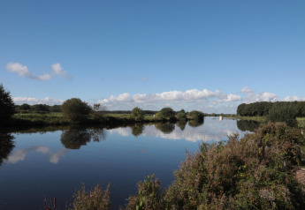 View of a serene lake with calm water, green foliage, and blue sky near a glamping accommodation.