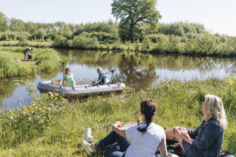 Vier mensen genieten van een zonnige glampingdag bij het water, twee in een boot en twee aan de oever.