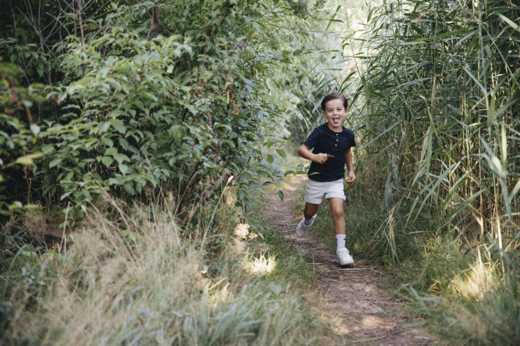 A happy boy runs along a leafy path surrounded by tall grass and bushes, photographed near a safari tent.