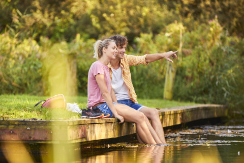 Stel zit ontspannen aan een steiger van een glampinglocatie met de voeten in het water.