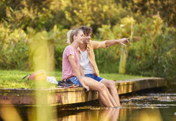 Pareja se relaja en un muelle de glamping con los pies en el agua, disfrutando de la naturaleza.