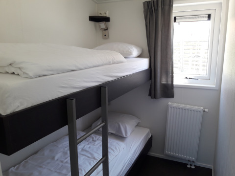 Kleine kamer met stapelbedden, raam met grijs gordijn en radiator in Premium Lodge bij Kompas Camping - Westende, België.