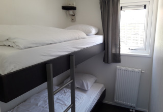 Habitación pequeña con literas, ventana con cortina gris y radiador en Premium Lodge de Kompas Camping - Westende, Bélgica.