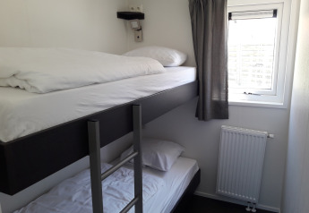 Kleines Zimmer mit Etagenbett, Fenster mit grauem Vorhang und Heizung im Premium Lodge von Kompas Camping - Westende, Belgien.