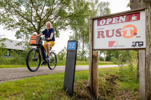 Un hombre sonriente pasa en bicicleta junto a un cartel de 'RUST' cerca de la Luxe belltent en CharmeCamping De Regge-Vallei.