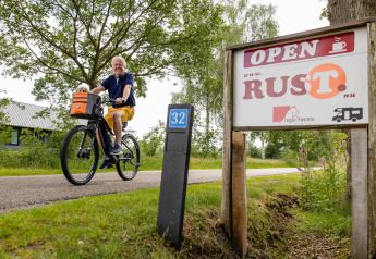 Un homme souriant fait du vélo près d’un panneau 'RUST' à la Luxe belltent du CharmeCamping De Regge-Vallei.