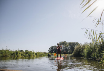 Personer på stand up paddle boards nyder naturen ved en flod tæt på et glampingophold i solen.
