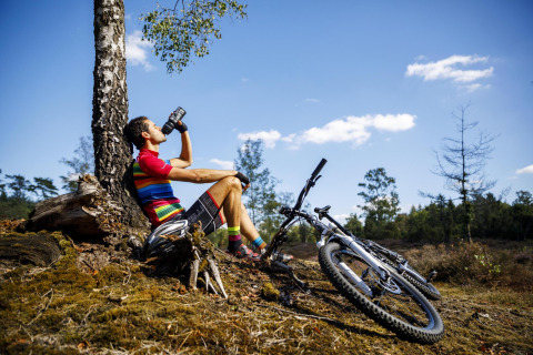 Ciclista con camiseta colorida descansando y bebiendo agua junto a su bici en Luxe belltent de CharmeCamping De Regge-Vallei.