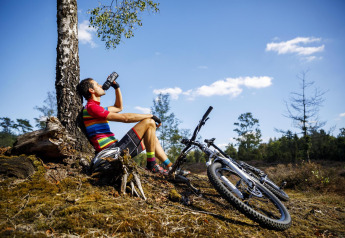 Cyklist i farverig trøje nyder en pause og drikker vand ved sit telt ved CharmeCamping De Regge-Vallei.