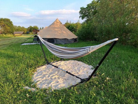 Safaritent en hangmat op een frame boven zand, in een grasveld met bomen en hutjes op de achtergrond.