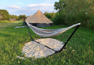 Safaritent en hangmat op een frame boven zand, in een grasveld met bomen en hutjes op de achtergrond.