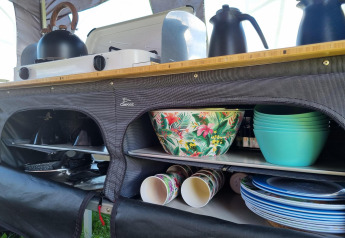 Ciotole colorate e stoviglie ordinate in una cucina da tenda safari con fornello e bollitore sopra.