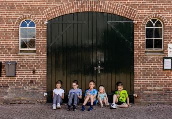 Fünf Kinder sitzen vor einem großen schwarzen Scheunentor bei Glamping Belltent, CharmeCamping De Regge-Vallei.