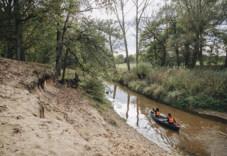 Zwei Personen fahren in einem Kanu auf einem ruhigen Fluss neben einem sandigen Ufer in einem Waldgebiet.
