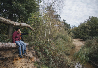 Two people sit on a tree trunk, looking out over a river and forest near a safari tent in the Netherlands.