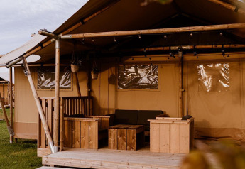 Ein gemütliches Glamping-Zelt mit Holzterrasse und rustikalen Möbeln steht auf einer grünen Wiese.