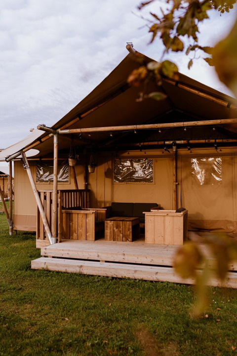 Tente de glamping extérieure avec terrasse en bois, mobilier rustique et pelouse sous un ciel nuageux.