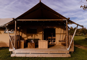 Foto di un alloggio glamping con tenda in tela, veranda in legno e mobili da esterno immersi nel verde.