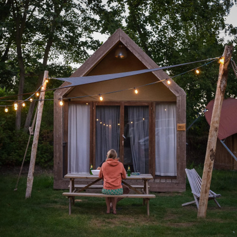 Person sitzt an einem Picknicktisch vor einer Boutique-Suite im Holiday Park Mölke, Niederlande, mit Lichterkette.