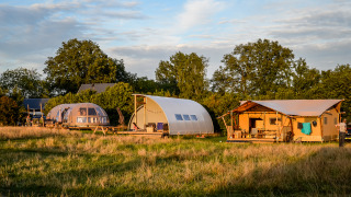 Tres tiendas de glamping modernas y coloridas se alzan sobre un campo de hierba, rodeadas de árboles y naturaleza.