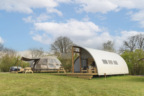 Two modern glamping lodges on a grassy field under a blue sky with clouds, surrounded by green trees.