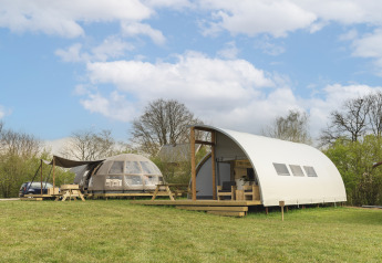 Two modern glamping lodges on a grassy field under a blue sky with clouds, surrounded by green trees.