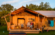 Et par sidder på verandaen foran et hyggeligt safaritelt på en campingplads omgivet af grønt.