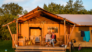 A couple sits on the porch of a cozy safari tent at a campsite, surrounded by trees and gear.