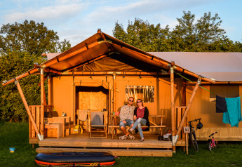Una pareja se sienta en el porche de una tienda de safari en un camping, rodeada de naturaleza verde.