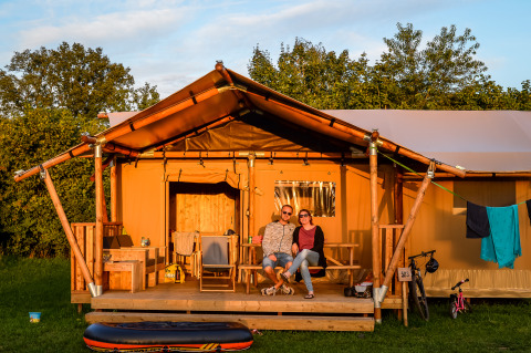 Un couple est assis sur la terrasse d'une tente safari confortable dans un camping entouré de verdure.
