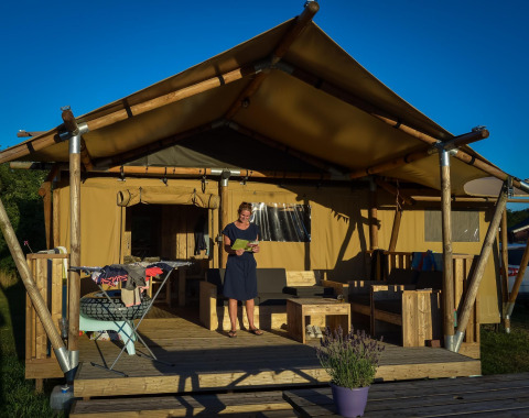 Eine Frau steht auf der Terrasse vor einem Safarizelt mit Holzmöbeln, Wäsche und Sonnenschein.