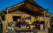 Una donna in piedi sulla veranda di una tenda safari con mobili in legno e bucato steso al sole.