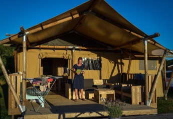 Een vrouw staat op het terras van een safaritent met houten meubels en wasgoed in de zon te drogen.
