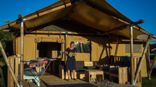 Een vrouw staat op het terras van een safaritent met houten meubels en wasgoed in de zon te drogen.